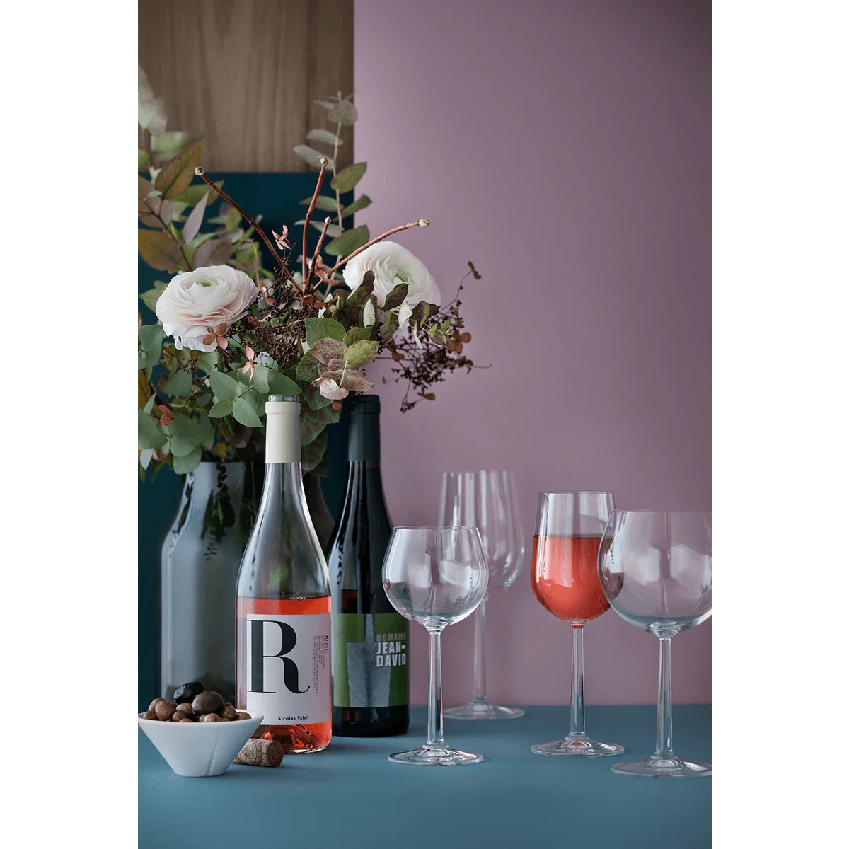 Sæt med ROSENDAHL Grand Cru vinglas med vin, flasker og blomster på en blå overflade.