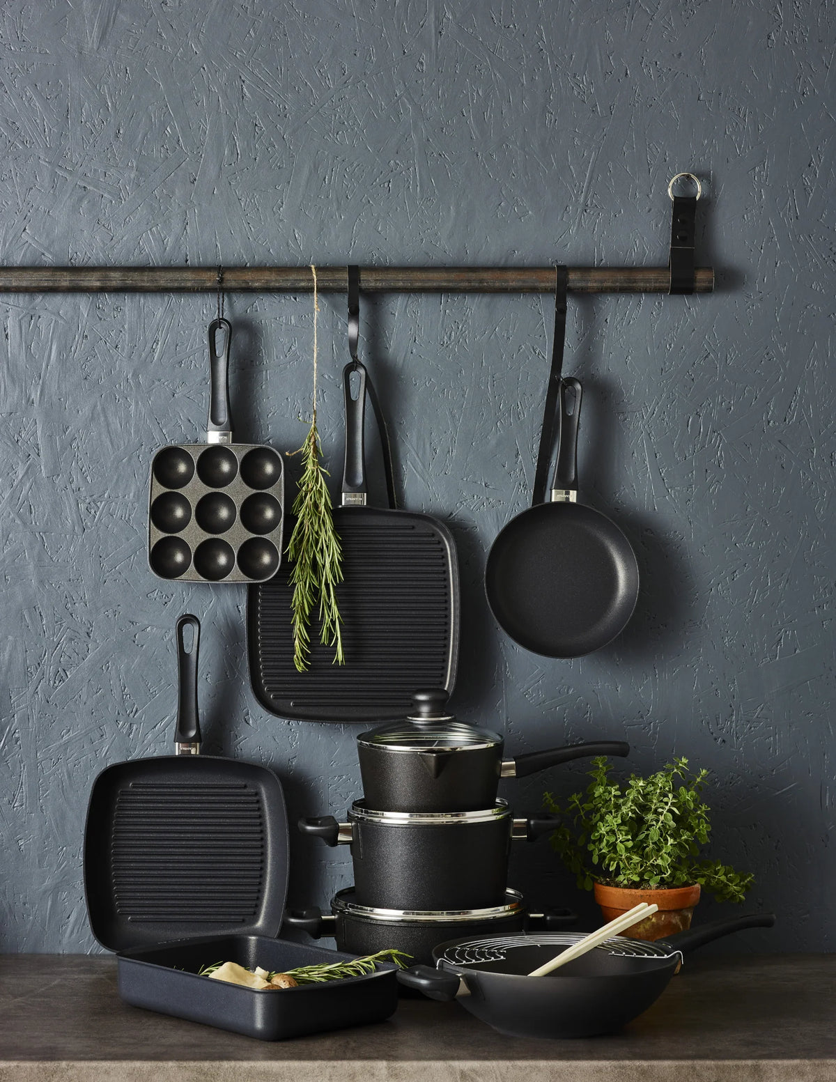 Sæt af sorte SCANPAN stegepander, grillpander og gryder på en mørkegrå væg.