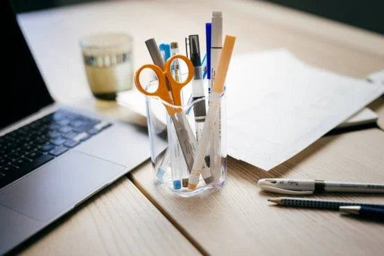 Orange Fiskars hobbysaks i en kop med skriveredskaber på et skrivebord ved siden af en laptop