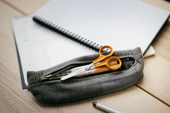Fiskars hobbysaks med orange håndtag i et gråt penalhus ved siden af en notesbog.