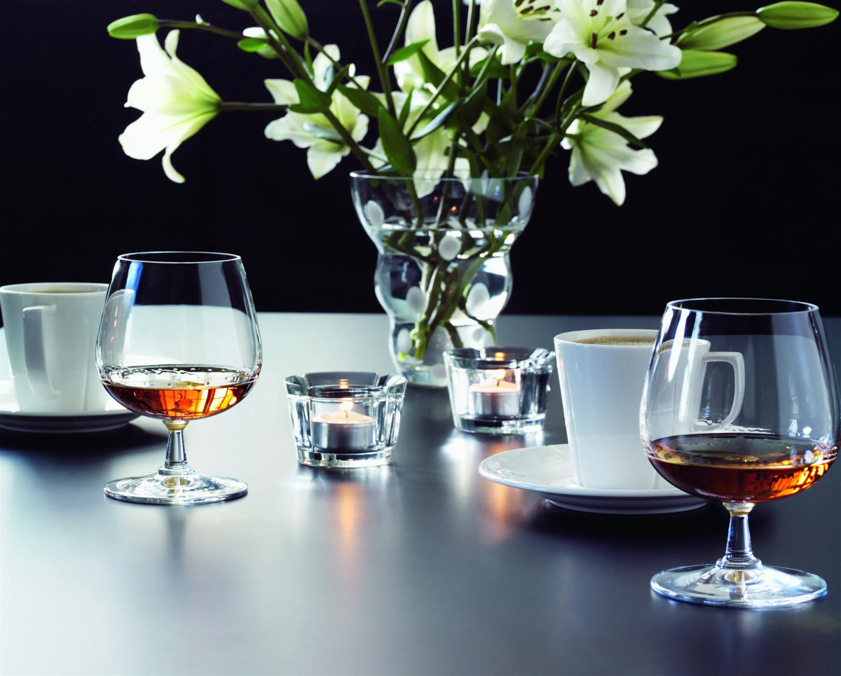 To ROSENDAHL Grand Cru cognacglas med cognac, kaffekopper og hvide liljer på et bord.