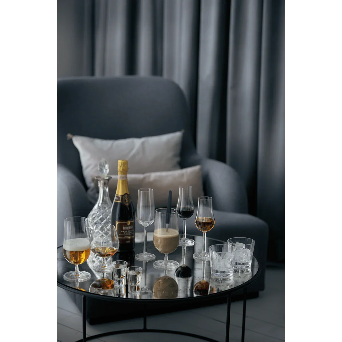 Sæt med ROSENDAHL Grand Cru glas og champagne på et lille rundt bord foran en grå lænestol.