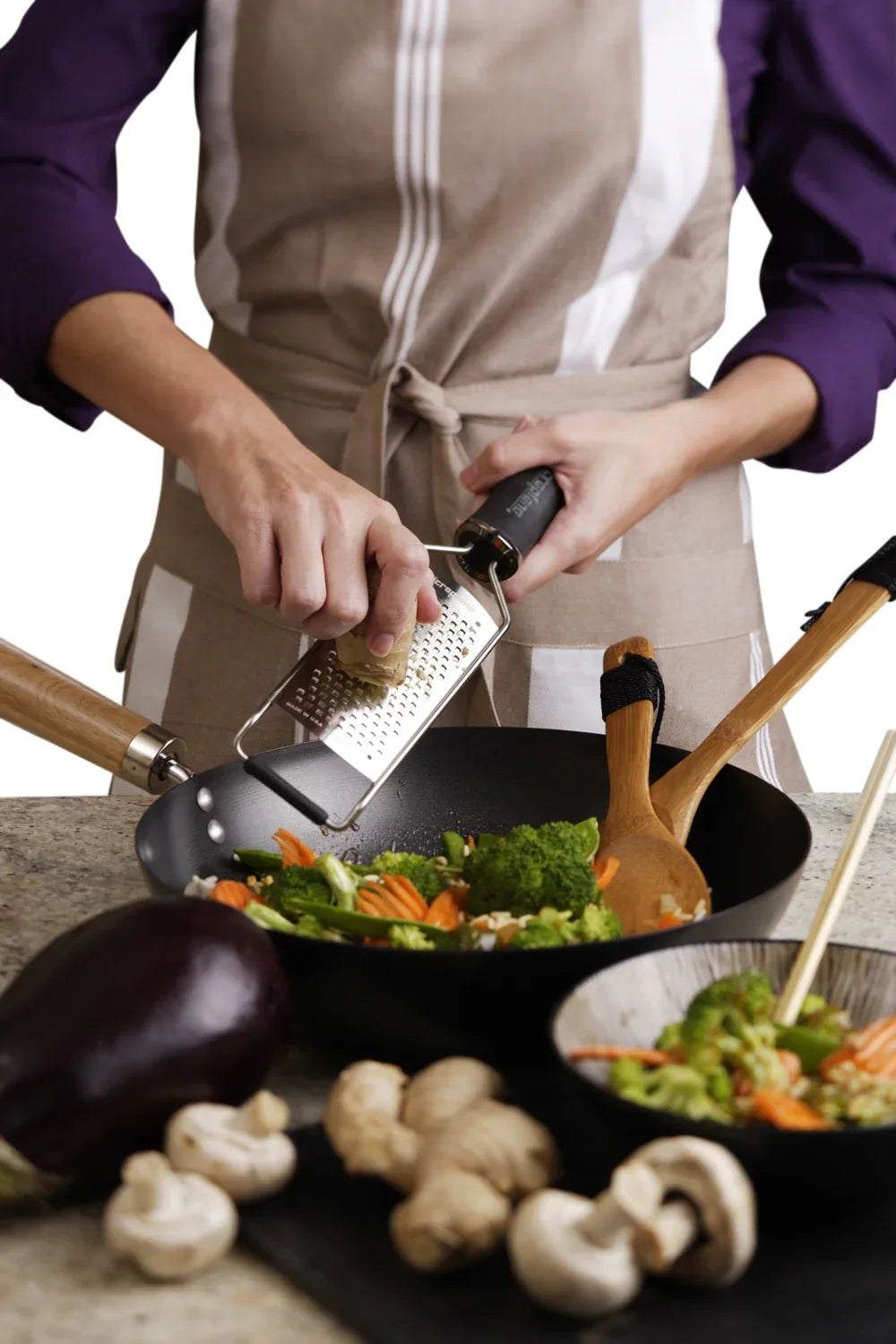 Person bruger et MICROPLANE rivejern til at rive ingredienser over en wok med grøntsager.