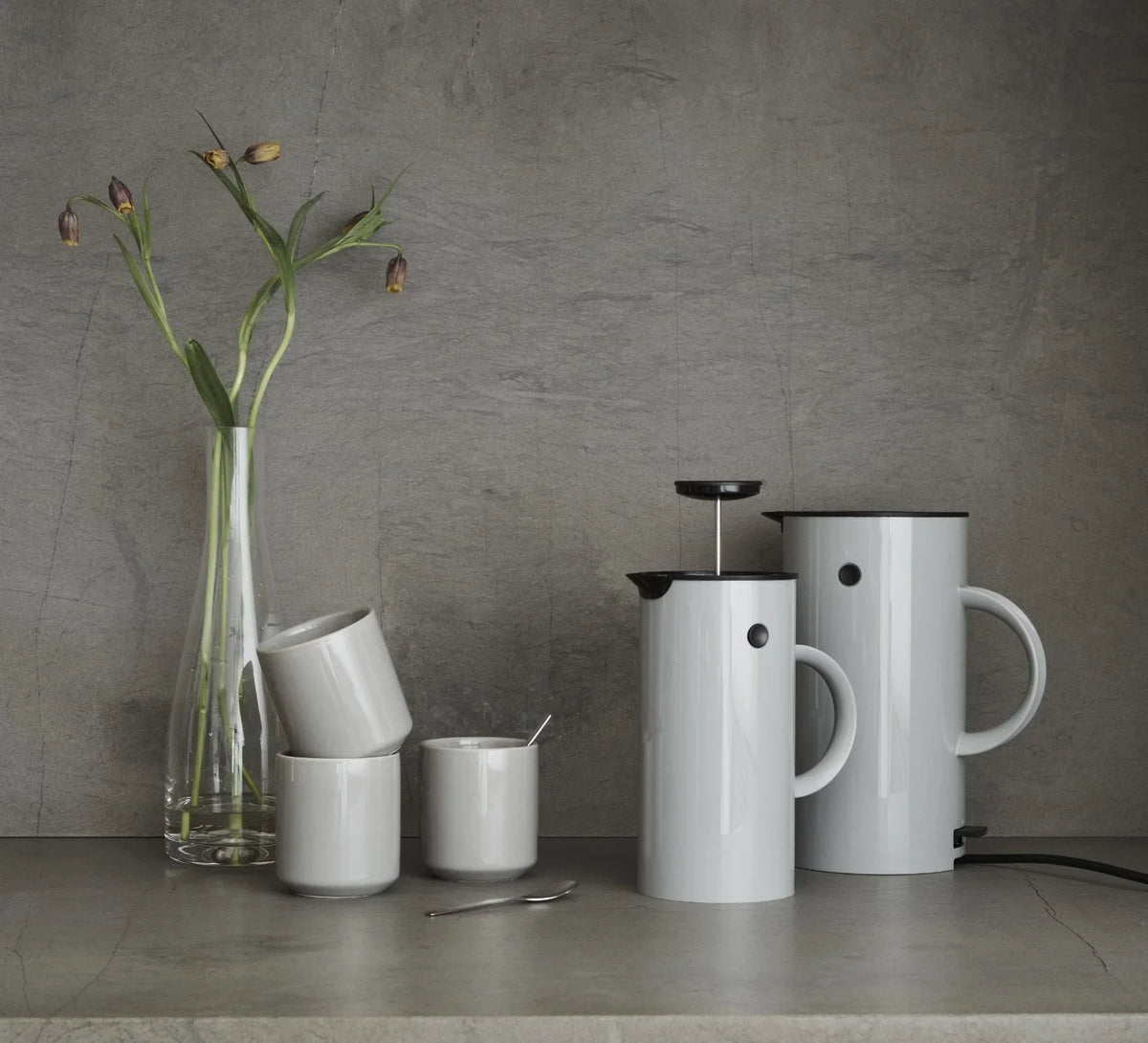Hvide STELTON stempelkander og kopper på en grå overflade med en vase med blomster.